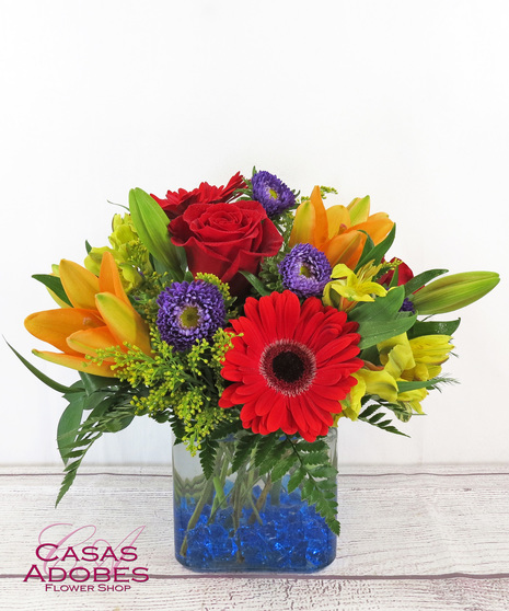 Cube vase with blue gems in the bottom filled with bright orange gerbera daisy, orange liily, purple aster, red roses, yellow alstroemeria, 