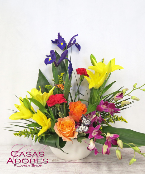  iris, yellow lilies, orange roses, red carnations, purple dendrobium orchids in a low garden bowl with delicate butterflies throughout the arrangement