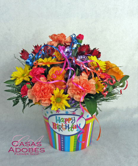 Pink carnations, orange roses, yellow daiy, pink lily in a bright and fun happy birthday container with streamers