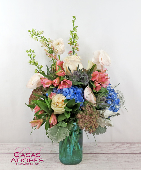 Roses, hydrangea, larkspur, ranunculus, thistle, and Queen Anne's lace presented in a colored glass vase