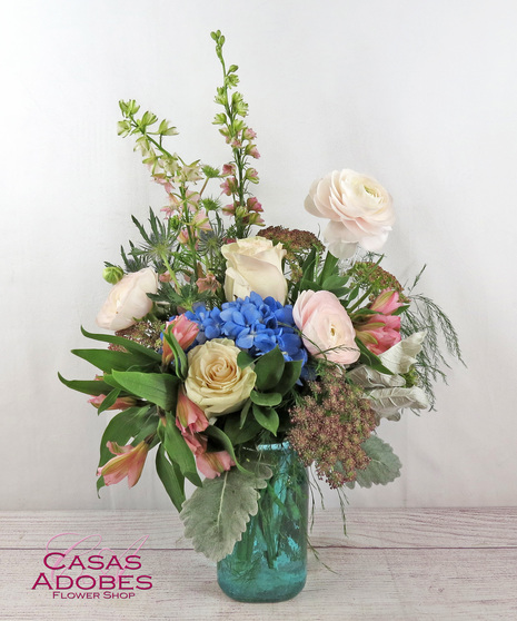 Roses, hydrangea, larkspur, ranunculus, thistle, and Queen Anne's lace presented in a colored glass vase