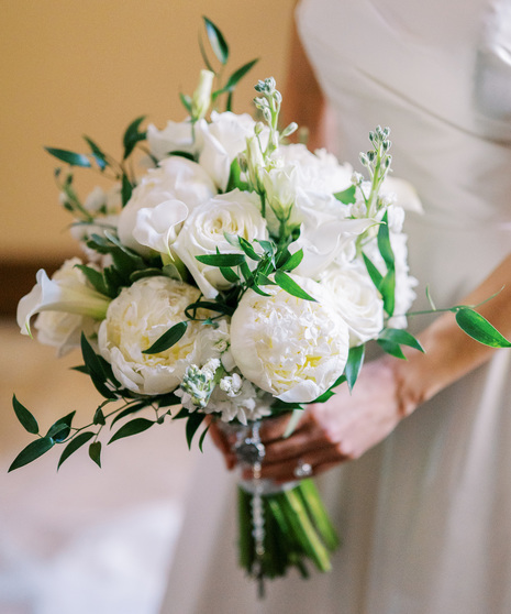 A loose hand-tied bouquet featuring gorgeous all-white roses, peonies, stock, miniature calla lilies and lisianthus with lush seasonal foliage.