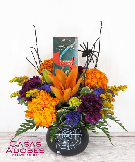  black cauldron with a spider web design, this floral bouquet overflows with fresh orange lilies, orange marigolds, deep purple carnations, solid aster and statice