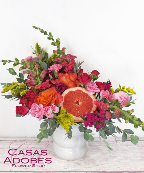 white vase with orange roses, hot pink snapdragons, yellow solidago, pink mini carnations and freshly sliced grapefruit