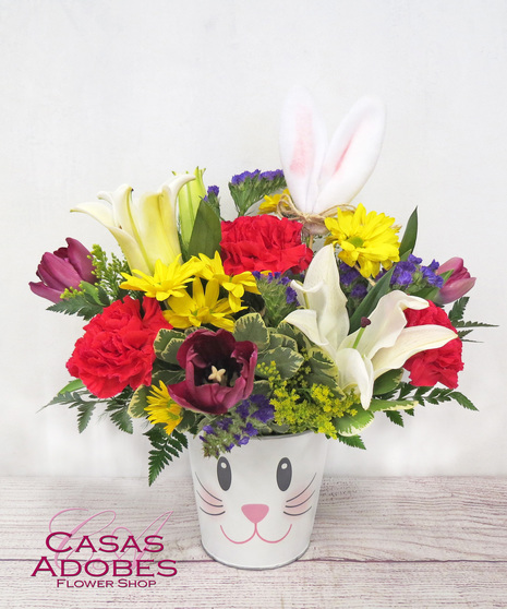 bunny bucket is filled with vibrant red carnations, yellow daisy chrysanthemums, purple statice, white stargazer lilies, and tulips